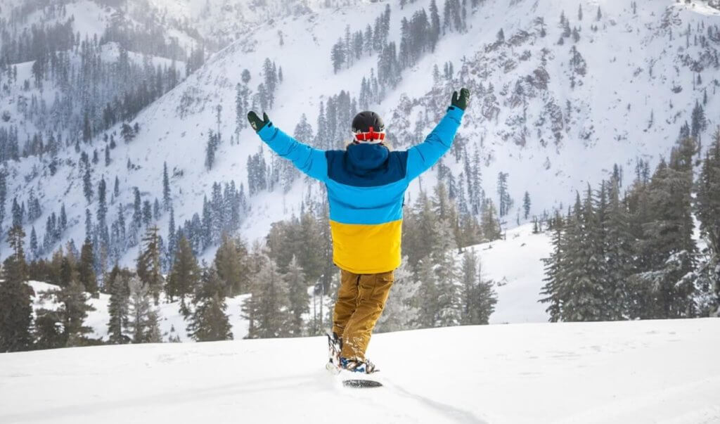 Snowboarder in fresh powder lifting arms in the air with snow-covered mountains in the background.