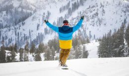 Snowboarder in fresh powder lifting arms in the air with snow-covered mountains in the background.