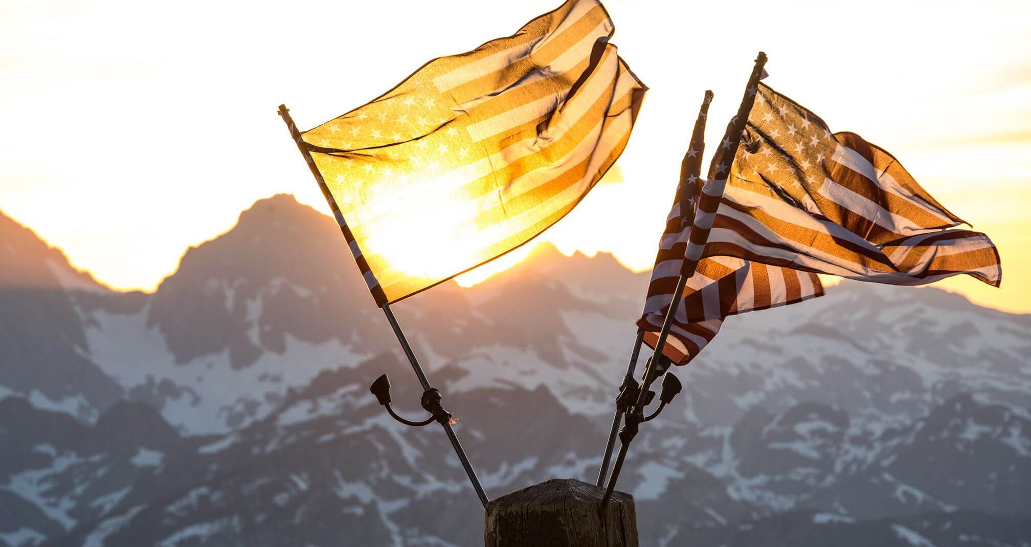 Three American flags flying in the breeze with the sun rising above a mountain range in the background