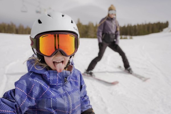 Little girl wearing a ski helmet and goggles sticking out her tongue, with an adult on skis blurry in the background