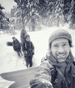 Professional snowboarder Jeremy Jones taking a selfie with his kids carrying their snowboards in the background walking through fresh snow.