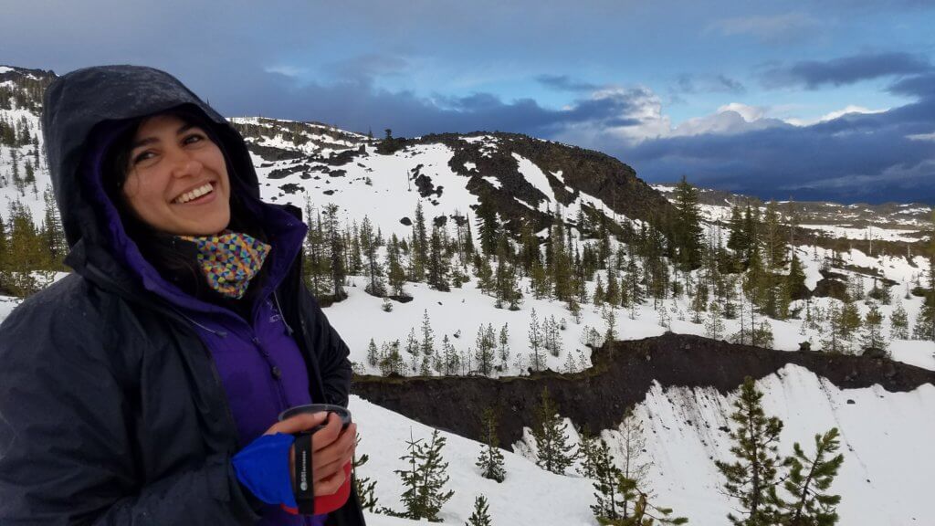 Photo of Elizabeth Sahagun, a BiPOC woman, bundled up in a winter coat with a snowy mountain landscape behind her.