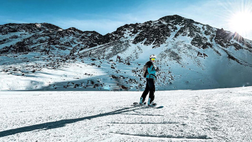 Woman on a snowboard with mountains in the background and a clear blue sky.