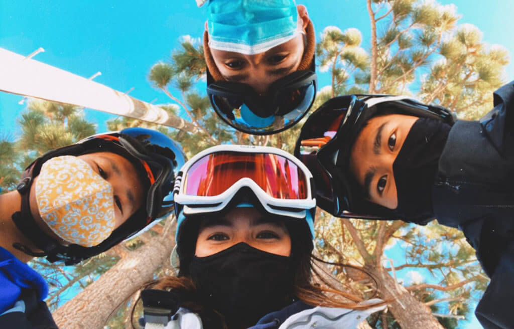 Four young people in ski gear and face masks circled together looking down at the camera