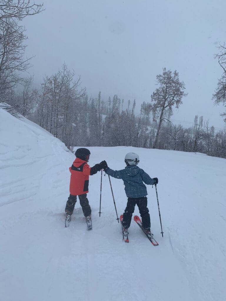 Two young boys in ski gear giving each other a high five on top of a snowy ski run during a Steamboat mom-son trip.