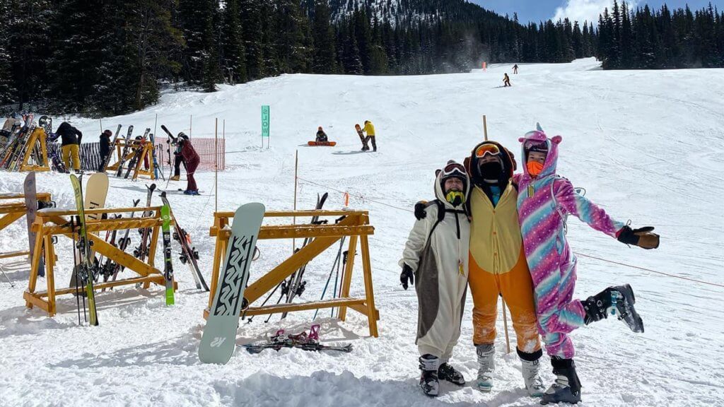 Three friends in colorful onesies and ski boots at the base of a ski resort