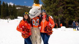 A horse mascot with an orange football jersey on stands between two young women in orange jackets holding orange and blue pom poms standing on a snow covered mountain.