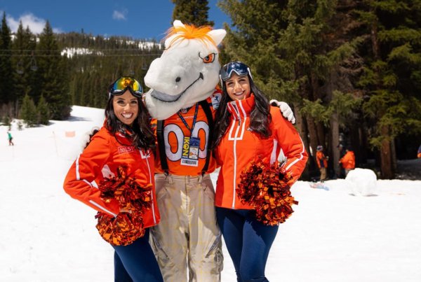 A horse mascot with an orange football jersey on stands between two young women in orange jackets holding orange and blue pom poms standing on a snow covered mountain.