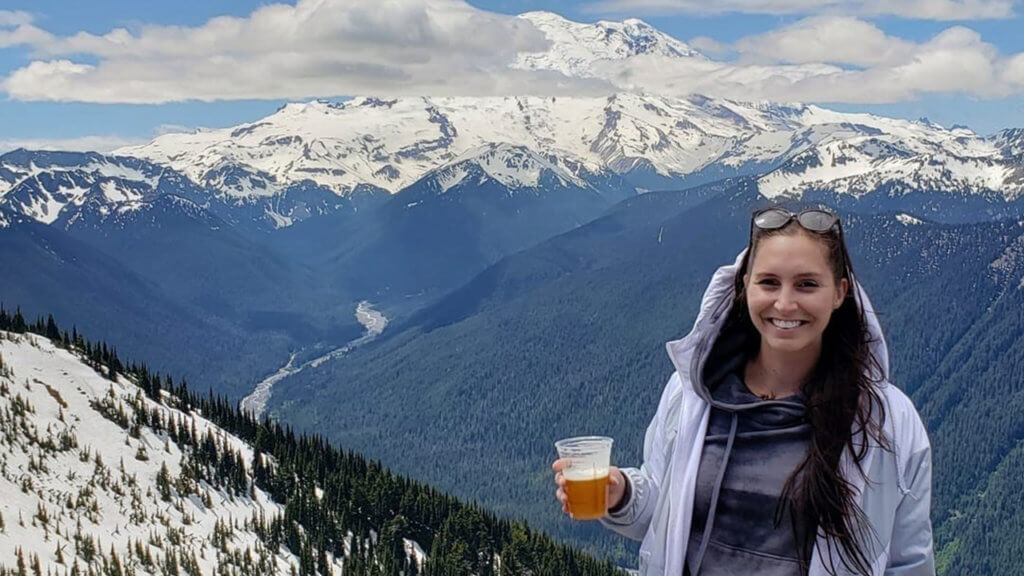 Caucasian girl at the top of a mountain holding a beer in front of Mt Rainier in Washington