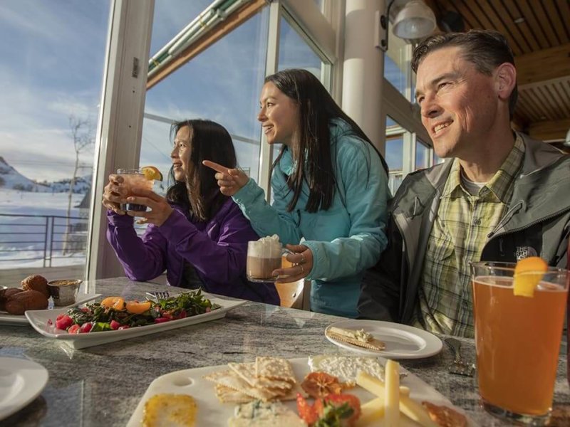 Three people (two women and one man) standing at a table covered in plates of food and drinks. The people are looking out the window at the mountain view.