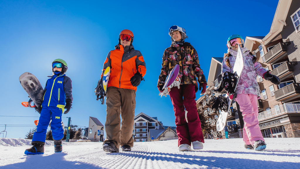 Four people (two adults, and two school-aged children) dressed in snow sports gear and each holding a snowboard, walking on groomed snow with a mountain lodge behind them on a blue sky day.