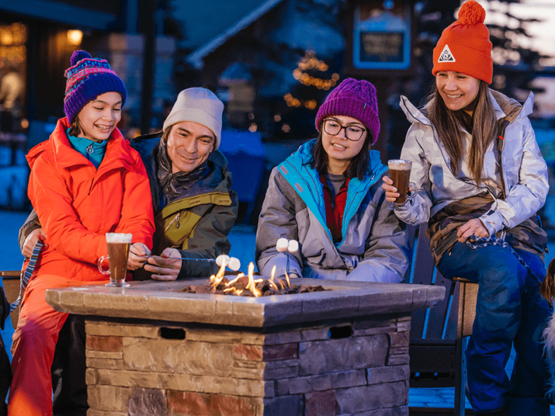 A group of people in winter gear and a dog sit next to a fire pit outside sipping warm drinks