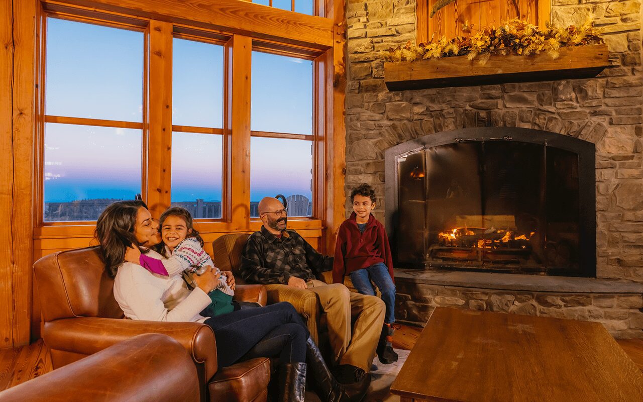 A family (two adults, and two children) sitting on a brown leather couch in a mountain cabin with a stone fireplace behind them with a blazing fire.