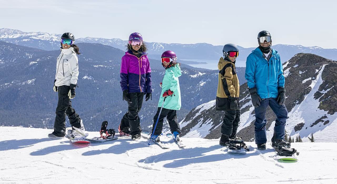 A family of 5 people turned towards the camera dressed in snow sport gear, buckled into snowboards and skis at the top of a ski run with views of mountains behind them.