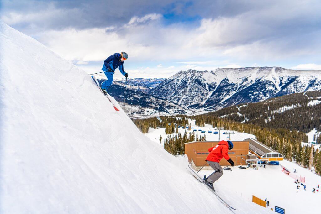 Two skiers racing down a steep snow slope with a scenic mountain backdrop behind them