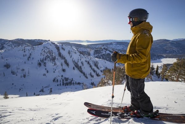 Skier in a yellow jacket standing at the top of a ski run looking out at a mountain landscape