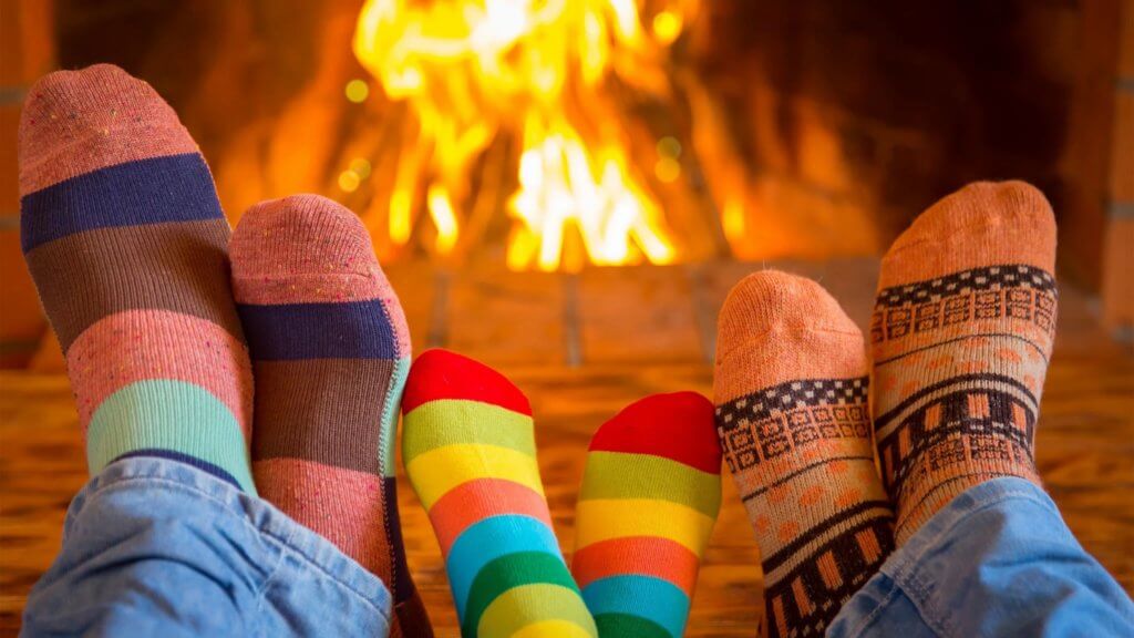 Three pairs of feet wearing colorful ski socks propped up in front of a fireplace. Ski socks should be an essential item on a ski trip packing list.