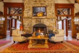 Two women sitting in comfortable chairs in front of a stone fireplace as they work on laptops