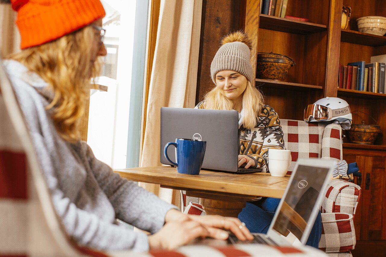 Two women sitting in a mountain lodge working on laptops and drinking coffee