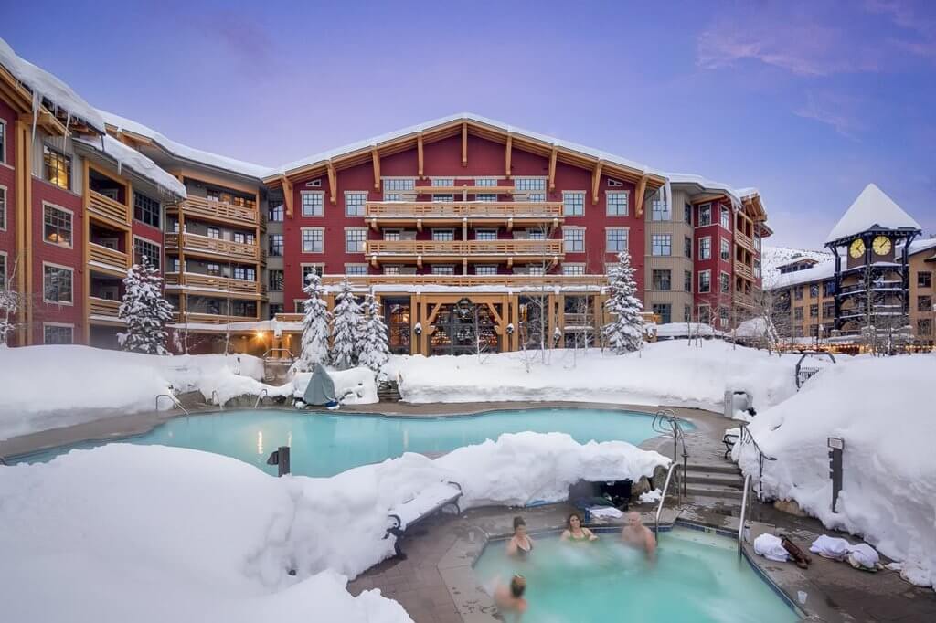 A heated pool and a hot tub surrounded by piles of snow and a red mountain lodge behind them
