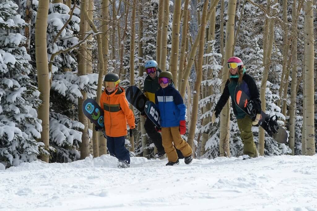 A family dressed in winter clothes carrying snowboards walking through snow
