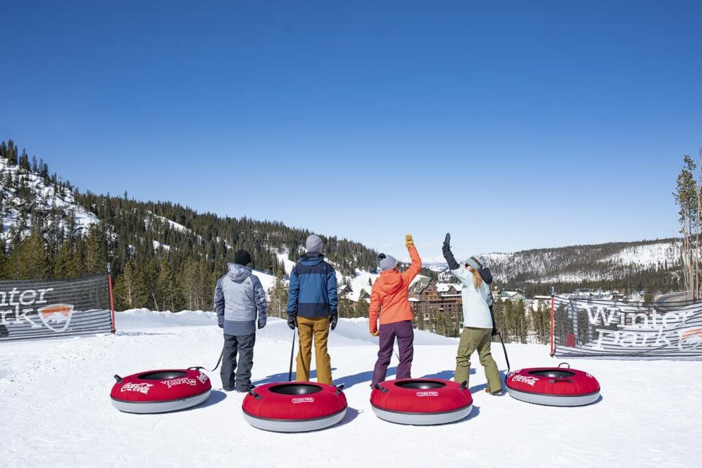 Four people dressed in winter snow gear standing at the top of a snowy hill with four inflatable tubes behind them
