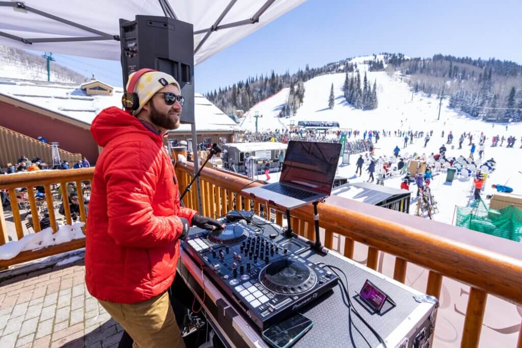 A DJ standing on a patio overlooking a crowd gathered at the base of a snowy ski hill.