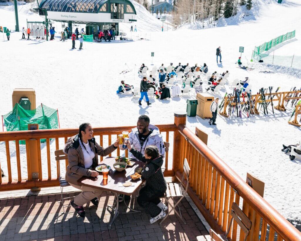 A family sitting at a table on a mountain lodge patio, overlooking a "snow" beach with people sitting in chairs on a sunny day