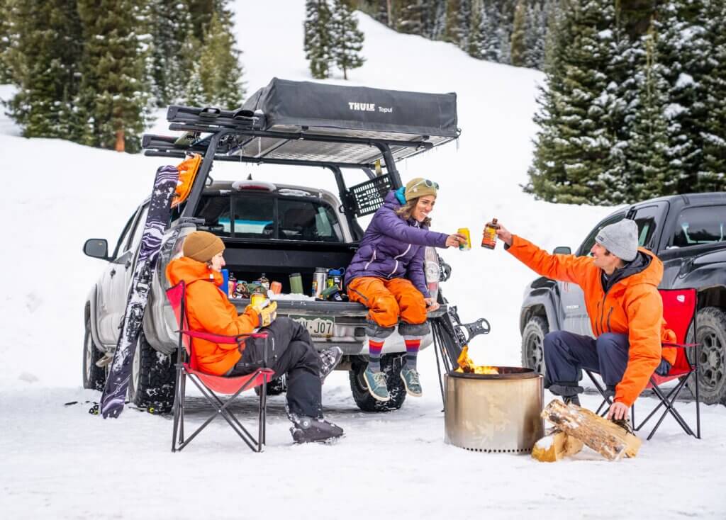 People dressed in ski gear sitting in folding chairs around a fire pit in a parking lot at the base of a ski resort