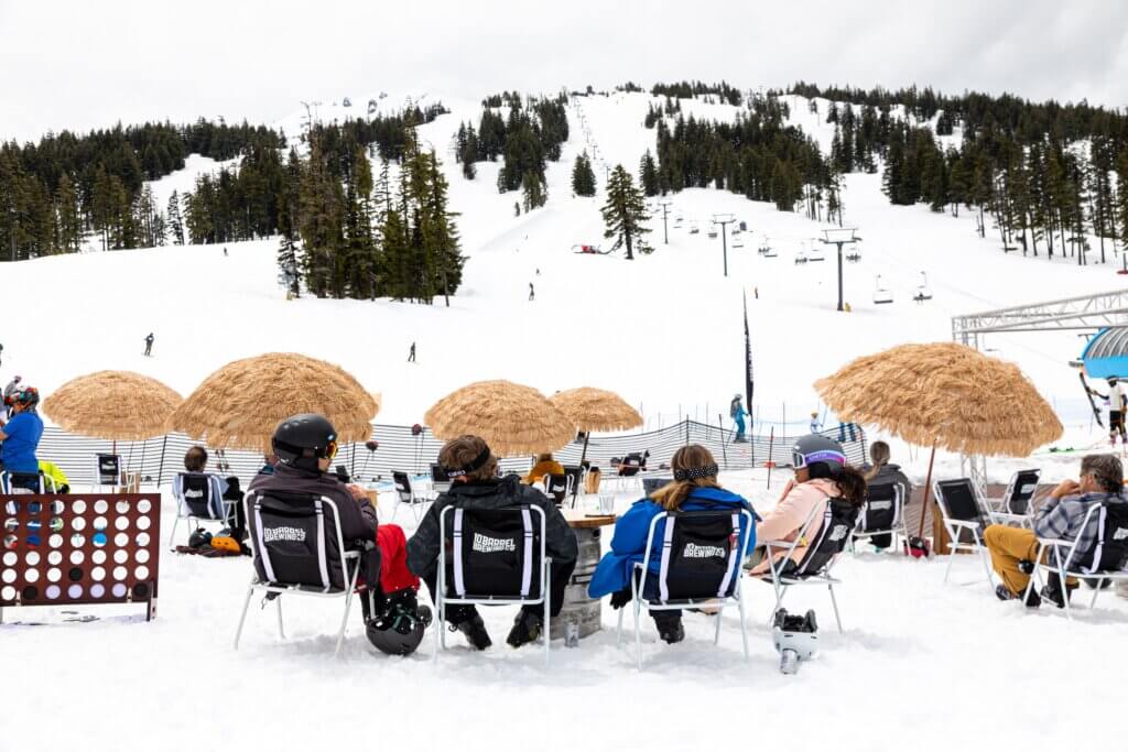 People sitting in folding chairs with straw umbrellas stuck into the snow at the bottom of a ski trail at one of the best mountain beach scenes
