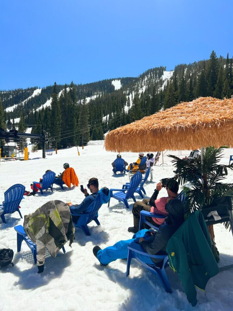 People sitting in chairs under straw umbrellas stuck into the snow at the bottom of a ski hill