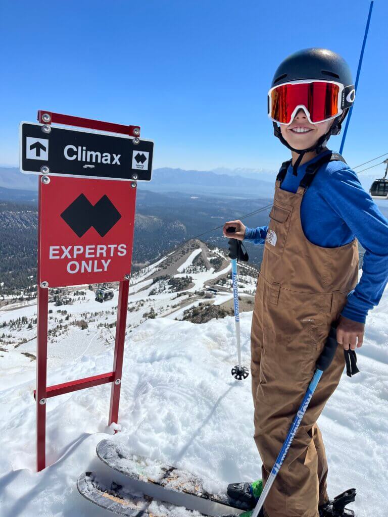 Young kid in ski gear standing at the top of a ski run, with a ski trail sign that reads "Climax. Experts Only"
