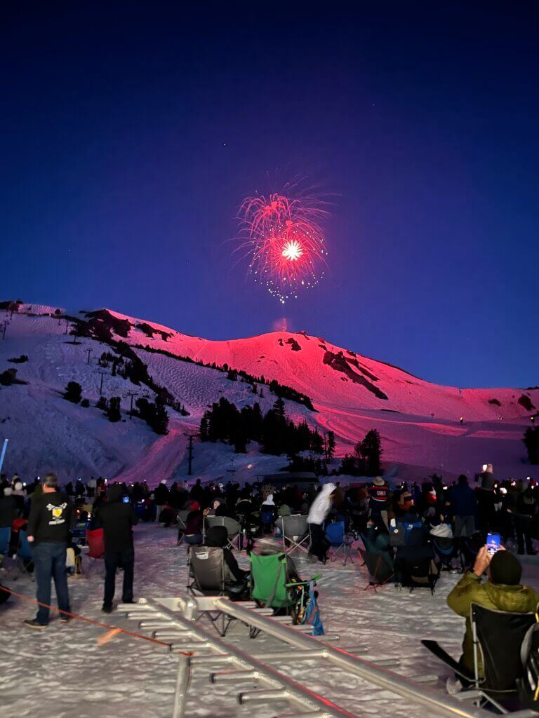 Crowd of people watching fireworks explode over a snowy mountain peak