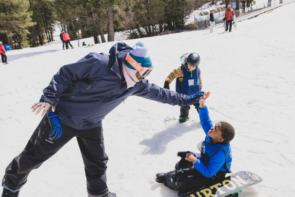 Adult high-fives a child who is taking a break from snowboarding.