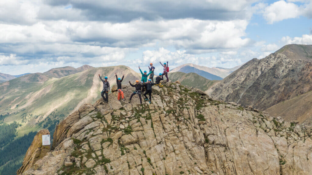 A group of climbers celebrating at the summit after completing a via ferrata ascent, with expansive mountain views in the background.