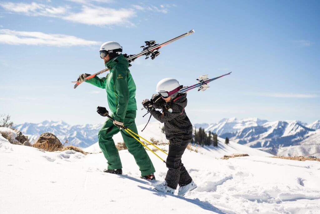 Ski instructor and child walk up a hill while holding ski equipment.