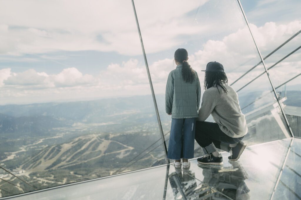 Two people standing on a glass floor looking out over the mountains