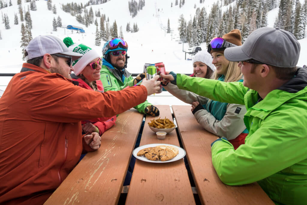 A reunion of people in winter gear sitting at a picnic table outside, toasting with their drinks, with a snowy mountain scene behind them
