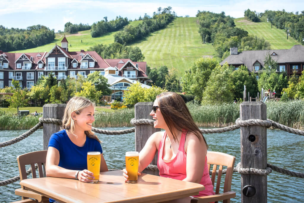 Two people sitting at a table holding glasses of beer as they sit on a deck next to a mountain lake