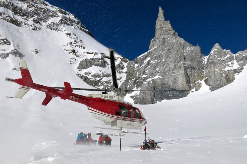 A helicopter lifting off of a snow-covered mountain, with people watching as it takes off