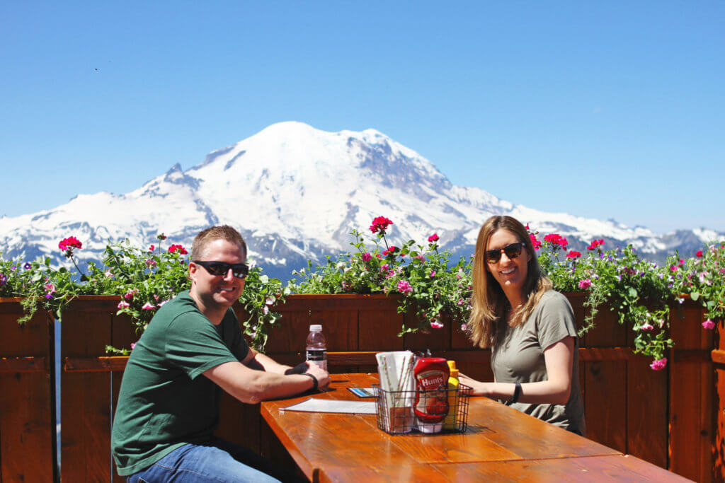 Two people smiling at the camera as they sit at a outdoor dining table with a snow-covered mountain in the background