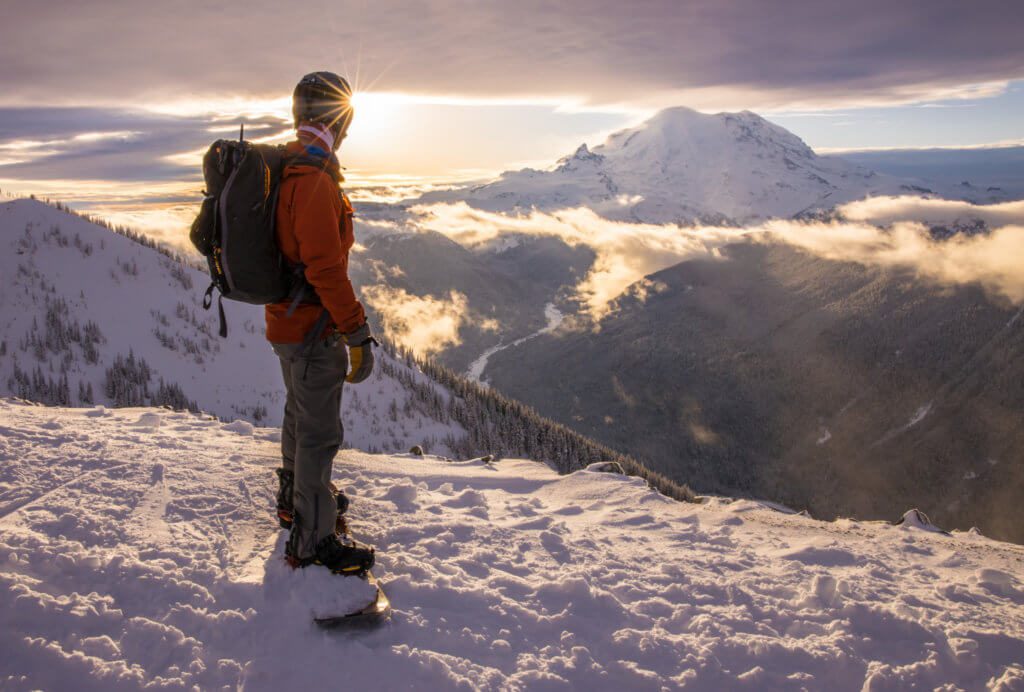 Snowboarder standing on top of a run at Crystal Mountain looking at Mt. Rainier in the background during sunset