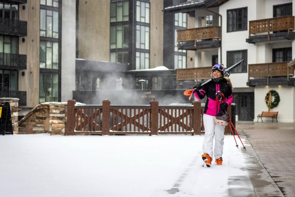 Person in winter gear holding skis over their shoulder walking in front of a ski lodge