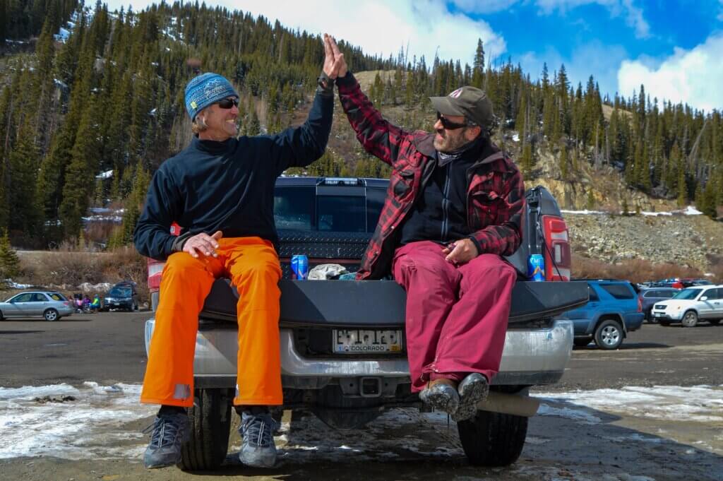 Two people sitting on the back of a truck, giving each other a high five, as they hang in the parking lot at Arapahoe Basin ski area