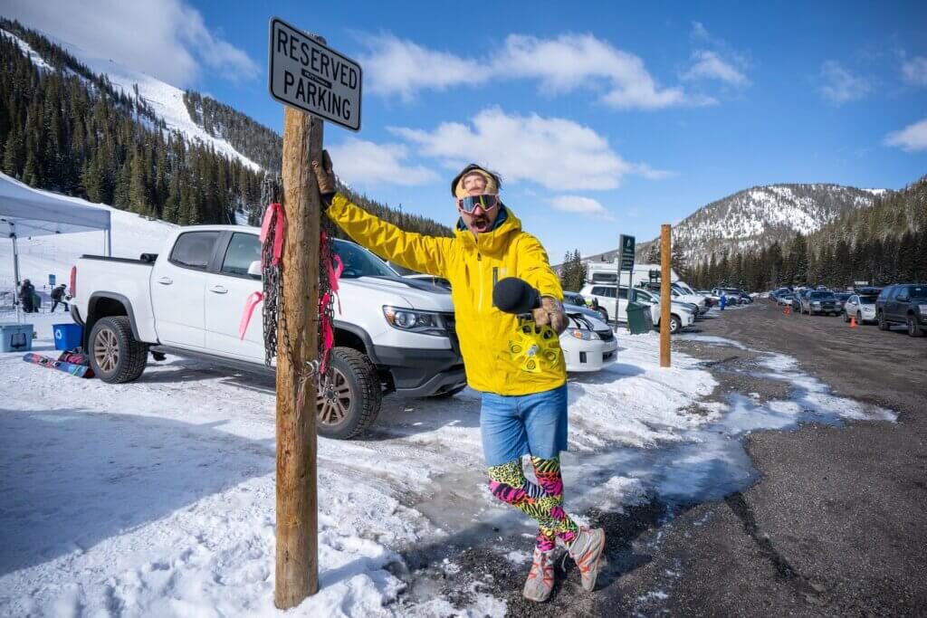 Someone in winter gear leaning against a sign in a snowy parking lot at the base of a ski resort