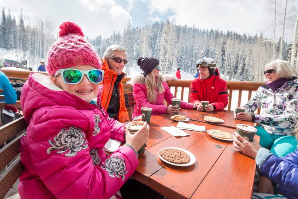 A young girl dressed in pink winter gear sitting at a table with adults. The girl is drinking hot chocolate and looking at the camera as the adults converse behind her, with a mountain scene in the background.